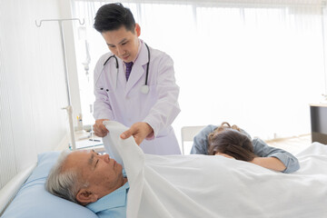 A young asian male doctor wearing white lab coat and stethoscope standing next to patient bed use...
