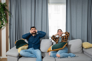 Young couple is sitting on a sofa in their apartment looking up and holding their hands to plug...
