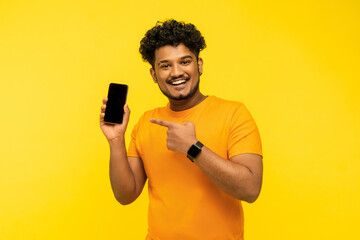 Fototapeta na wymiar Smiling Indian guy on a yellow background holds a smartphone with a black screen in his hand, points to it with his hand, looks at the camera