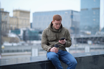 Obraz premium young man using smartphone and listens to music in bluetooth headphones. lonely man in autumn jacket drinks coffee while walking in the city