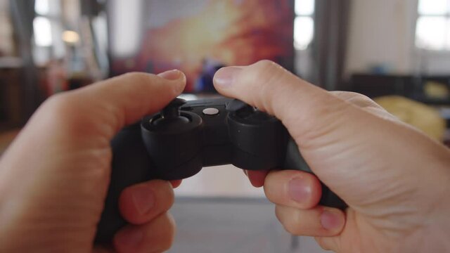POV of man moving joysticks and pressing buttons on black console controller while playing shooter video game on TV at home