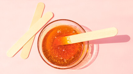 Shugaring paste in a glass bowl on a pink background. Hair removal, epilation, wooden spatulas for sugaring paste.