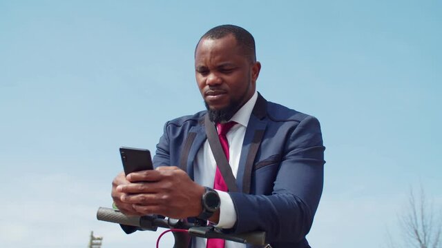 Portrait of successful confident handsome african american businessman in formal wear leaning on steering wheel of electric kick scooter, networking online and text messaging on cellphone outdoors.