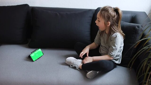 Little caucasian girl sitting on couch at home making speech therapy exercises looking in phone screen, mock up with green screen