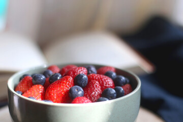 Bowl of blueberries and strawberries on wooden tray and open book. Selective focus, dark textile background.