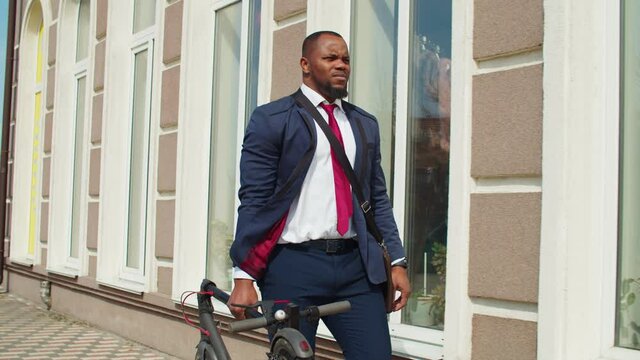 Confident good looking bearded african american male entrepreneur in trendy suit carrying folded electric scooter, going to work while walking along sidewalk in city on spring sunny day.