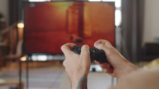 Over the shoulder close up shot of hands of unrecognizable man using wireless controller while playing shooter video game on TV at home