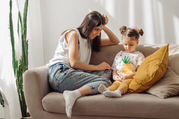 A dark-haired young mixed-race woman plays on the couch with her little daughter.Home interior design with houseplants.Time together.Family concept and Mother's Day concept.