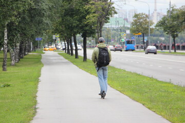 Russian man with backpack drive on compact electrical kick scooter on right side of asphalt street road alley on summer day, rear view