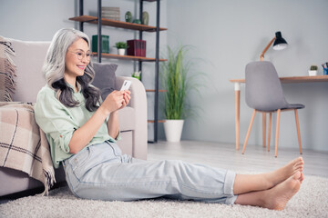 Full length body size side profile photo of senior business woman typing message on cellphone sitting barefoot on floor