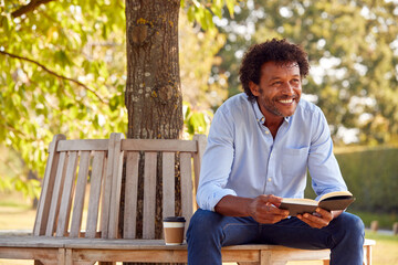 Mature Man Relaxing Sitting On Park Bench Under Tree Reading Book With Takeaway Coffee