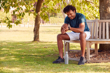 Man Wearing Fitness Clothing Sitting On Seat Under Tree Checking Activity Monitor On Smartwatch