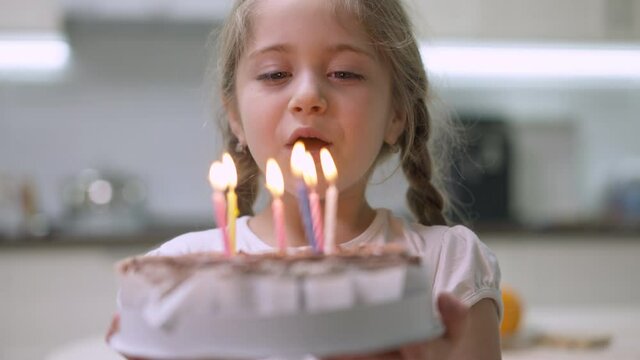 Close-up of charming little girl with closed eyes making wish and blowing candles on birthday cake. Portrait of happy Caucasian pretty child celebrating birthday indoors at home. Joy and lifestyle