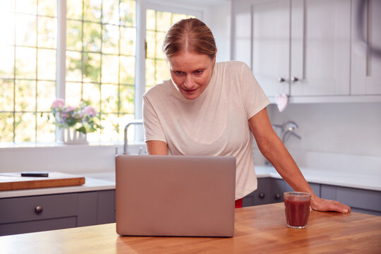 Mature Woman Wearing Fitness Clothing At Home In Kitchen Logging Activity On Laptop