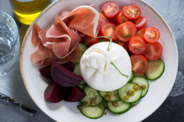 Closeup of a beige plate with burrata cheese, prosciutto ham, beetroot, cucumber and cherry tomatoes, elevated view
