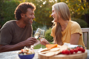 Mature Couple Celebrating With Champagne As They Sit At Table In Garden With Snacks