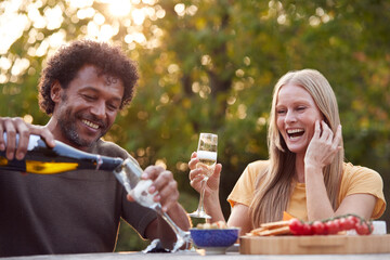 Mature Man Pouring Champagne As Couple Celebrate Sitting At Table In Garden With Snacks