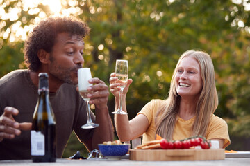 Mature Couple Celebrating With Champagne As They Sit At Table In Garden With Snacks
