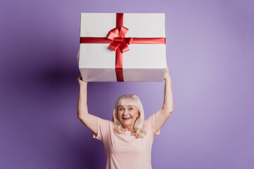 Amazed old woman hold get big gift box package isolated over purple background