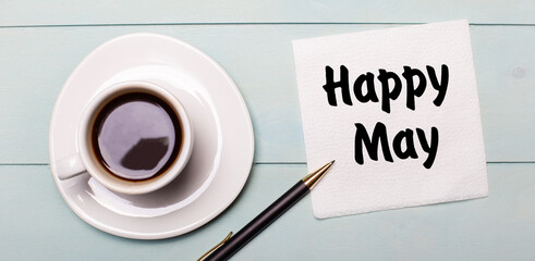 On a light blue wooden tray, there is a white cup of coffee, a handle and a napkin that says HAPPY MAY