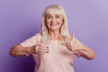 Photo of positive old woman hold water glass show thumbup sign on purple background