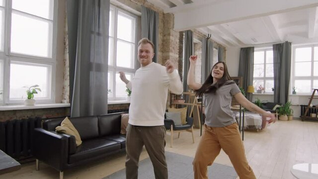 Dolly-in shot of young man and woman looking at camera and smiling while dancing together in their loft apartment