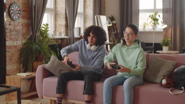 Cheerful teenage boy and girl sitting together on sofa in living room, smiling and using controllers while playing console game