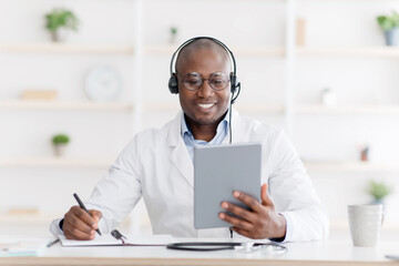 Online work with patients. Cheerful african american doctor in headphones holding digital tablet in...