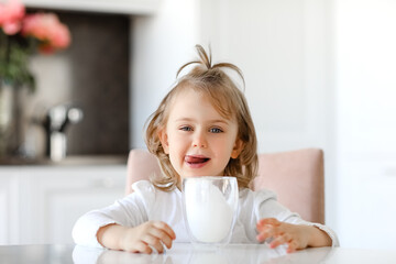 Blond cute child girl with blue eyes with mustache of milk on the lips is holding a glass of milk...