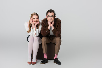 happy couple in retro clothes looking at camera while sitting on suitcase on grey background