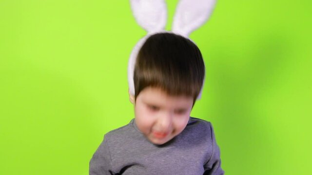 A happy funny boy in rabbit ears on his head tries to throw his ears off his head, nods his head, plays and laughs against a yellow background. Easter children