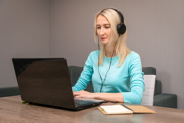 Young blonde woman sitting at a desk in large headphones at a laptop, listening to a lecture or webinar, learning via the Internet, using a PC, improving language skills