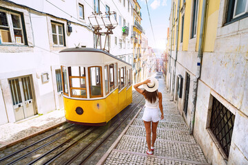 Woman tourist walking in narrow streets of Lisbon city old town. Famous retro yellow funicular tram on a sunny summer day. Tourist attraction. Vacation and travel concept. - obrazy, fototapety, plakaty