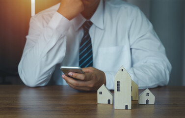 Business  Finance concept. Close up wooden house model with young agent businessman using mobile phone ongoing to calculate home loan mortgage to summary expense payment. Technology Insurance housing.