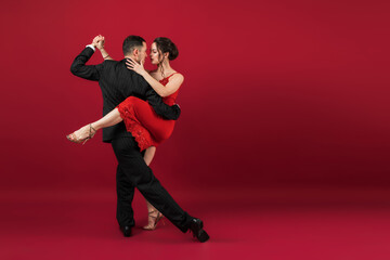 Fototapeta na wymiar Couple of professional tango dancers in elegant suit and dress pose in a dancing movement on red background. Handsome man and woman dance looking eye to eye.
