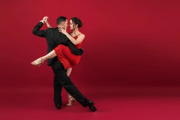 Fotobehang Couple of professional tango dancers in elegant suit and dress pose in a dancing movement on red background. Handsome man and woman dance looking  eye to eye. © Artem Bruk