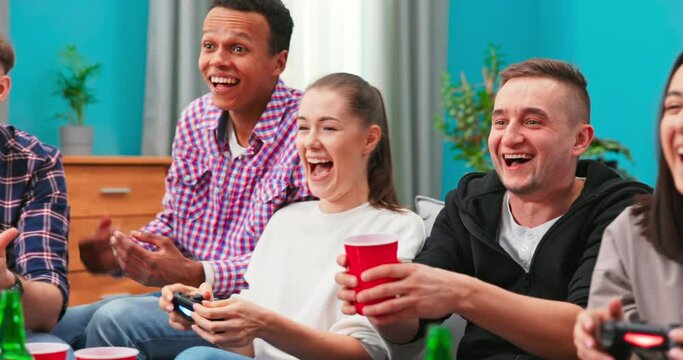 Multinational friends sitting at home on couch and playing video games on console. Cheerful mixed races friends resting in room sitting on sofa. A group of college students are at a dorm party.
