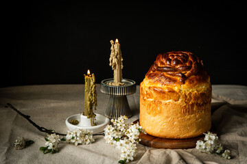 Homemade Ukrainian traditional Easter bread -- paska, natural bee wax candles and white spring flowers on linen tablecloth on dark background.