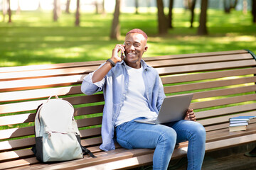 Fototapeta premium E-learning concept. Funky black student with laptop talking on smartphone on bench at park