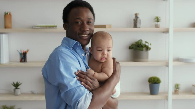 Cheerful african american dad holding and lullying his adorable newborn baby, smiling at camera at living room interior