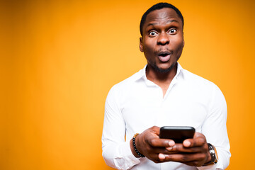 Handsome african american man holding a phone in his hand and is surprised by its power while standing on yellow background