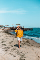 Adult blond woman with straw summer hat and yellow cotton shirt walks along the beach on a sunny day