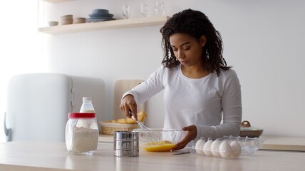 Young african american lady preparing dough for pancakes, whisking eggs and pouring milk, cooking...