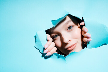 Smiling face of a young painted girl looking at the camera through a hole in a blue background revealing her hands