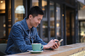 portrait of Asian young man at outdoor cafe, using mobile phone, with happy smilie