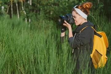 A girl in a gray jacket with a yellow backpack holds a professional photo-video camera in her hands. Against the background of green nature and forest.