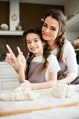 Happy mom and daughter in aprons cook in the kitchen and raise their index fingers in flour while looking at the camera