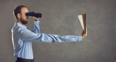 Profile view of serious young guy who needs glasses looking in binoculars and reading book he's...