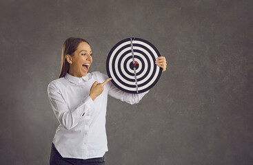 Happy excited smiling young woman holding dartboard target with red dart right in bullseye standing on gray studio background. Business goal, target market, objective, success concept