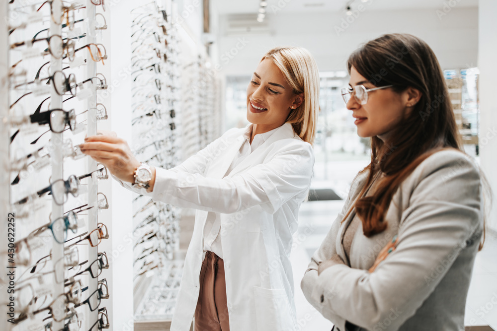 Poster beautiful and fashionable woman choosing eyeglasses frame in modern optical store. female seller spe